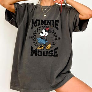 Może przedstawiać: Ciemnoszara koszulka z nadrukiem Minnie Mouse w stylu vintage. Projekt zawiera napis "MINNIE MOUSE" oraz lata "1928" i "1928". Myszka Minnie jest przedstawiona w swoim klasycznym stroju, z niebieską sp&oacute;dnicą i ż&oacute;łtymi butami.