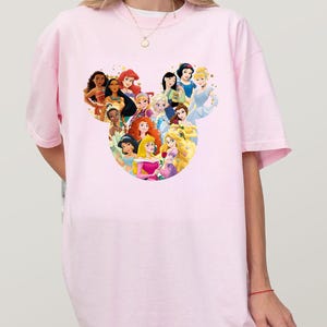 May include: A light pink t-shirt featuring a graphic of various Disney princesses arranged in a Mickey Mouse head shape. The princesses are depicted in their signature outfits and hairstyles. The shirt is a casual, oversized style.