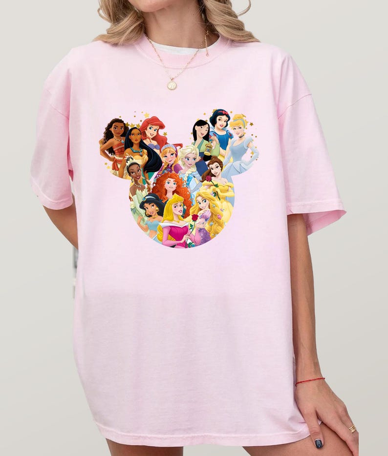May include: A light pink t-shirt featuring a graphic of various Disney princesses arranged in a Mickey Mouse head shape. The princesses are depicted in their signature outfits and hairstyles. The shirt is a casual, oversized style.