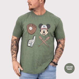 May include: Heather military green t-shirt featuring baseball-themed graphics. The design includes a baseball glove with a ball, Mickey Mouse wearing a baseball cap, a foam finger, and crossed baseball bats. The shirt is a comfortable, casual wear.