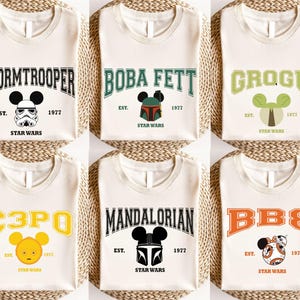 Op de afbeelding: Cr&egrave;mekleurige T-shirts met Star Wars-personages: Stormtrooper, Boba Fett, Grogu, C3PO, Mandalorian en BB8. Elk shirt heeft een personagenaam in een universiteitslettertype, een Mickey Mouse-hoofd en de tekst "EST. 1977 STAR WARS."