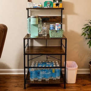 May include: A coffee bar setup with a light green Keurig coffee maker, various tea and coffee boxes, and a water bottle. The bar is on a dark brown and black metal shelving unit. The wall above has coffee-themed decorations.