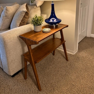 K&ouml;nnte beinhalten: Ein zweistufiger Beistelltisch aus Holz im Mid-Century-Stil. Auf der oberen Ebene steht eine dunkelblaue Lampe mit cremefarbenem Lampenschirm. Eine kleine Topfpflanze und Untersetzer befinden sich ebenfalls auf dem Tisch.