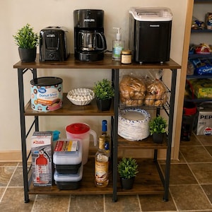 May include: A multi-tiered kitchen shelf with a black metal frame and brown wooden shelves. The top shelf holds a black toaster, coffee maker, and ice maker. Other shelves display food items, a fire extinguisher, and decorative plants.