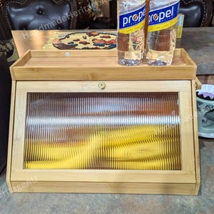May include: A light-colored bamboo bread box with a glass front and a small tray on top. The glass has a ribbed texture. Two water bottles are on the tray. A coffee maker is on the left.