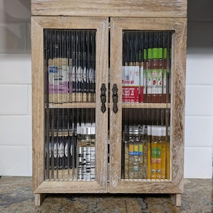 May include: A wooden cabinet with glass doors, featuring a variety of glass bottles and jars, some with labels. The cabinet has a distressed white finish and is ideal for storing and displaying small items.