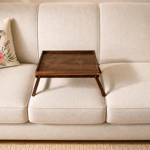 May include: A light beige sofa with a decorative pillow featuring a floral and bunny ear design. A dark brown wooden tray with folding legs sits on the sofa. The room has a neutral color palette with a rug and a plant.