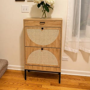 May include: A light wood cabinet with two drawers featuring woven panels and black hardware. The cabinet is set against a cream-colored wall with framed artwork and a vase of white flowers. The cabinet sits on black metal legs.