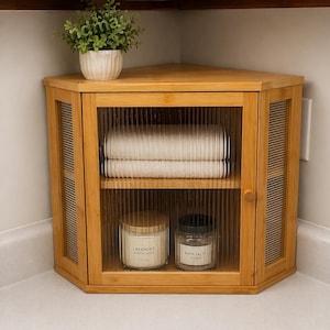 May include: A light brown corner cabinet with a glass door, holding folded white towels, a candle labeled "Lavender", and a jar of "Bath Salts". A small potted plant sits on top. The cabinet is made of bamboo.
