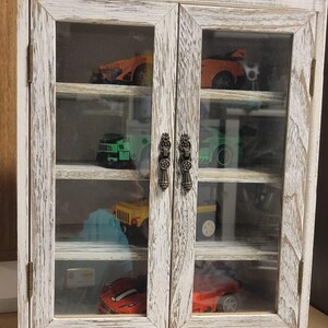 May include: A distressed white wooden display cabinet with glass doors, showcasing a collection of toy cars. The cabinet has two doors with ornate metal handles and multiple shelves. The toy cars are various colors.