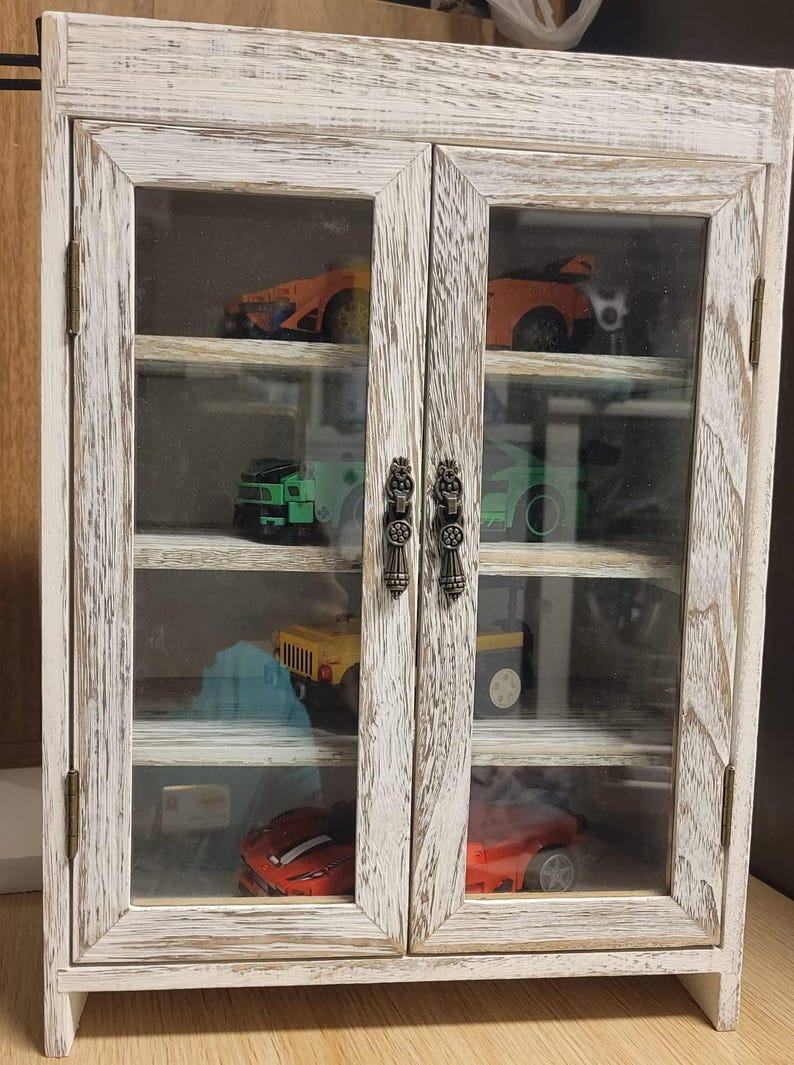 May include: A distressed white wooden display cabinet with glass doors, showcasing a collection of toy cars. The cabinet has two doors with ornate metal handles and multiple shelves. The toy cars are various colors.