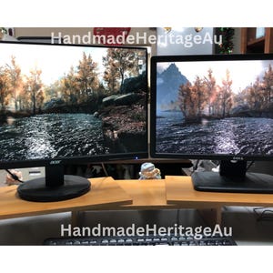 May include: Two computer monitors displaying a scenic landscape are placed on a light brown wooden monitor stand. The stand has a keyboard shelf. The text "HandmadeHeritageAu" is visible on the image.