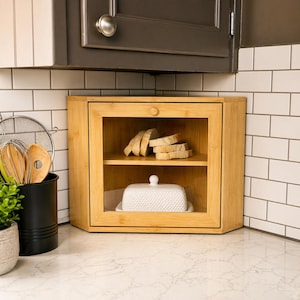 May include: A bamboo bread box with a clear glass door and a small wooden knob. Inside, there are slices of bread on a shelf and a white butter dish. The box is on a kitchen counter.