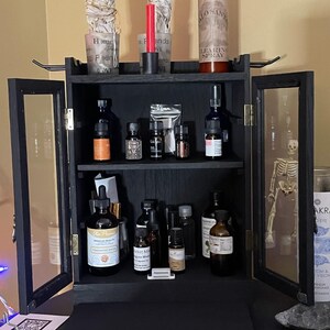 May include: A black wooden cabinet with glass doors, filled with various essential oil bottles, incense, and a bottle of "LEARING SPRAY". The cabinet is decorated with sage bundles, candles, and a small skeleton. The cabinet is on a black surface.