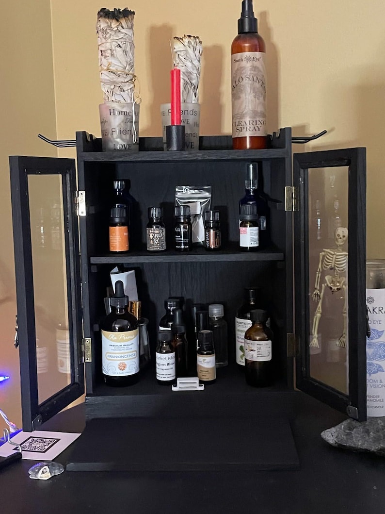 May include: A black wooden cabinet with glass doors, filled with various essential oil bottles, incense, and a bottle of "LEARING SPRAY". The cabinet is decorated with sage bundles, candles, and a small skeleton. The cabinet is on a black surface.