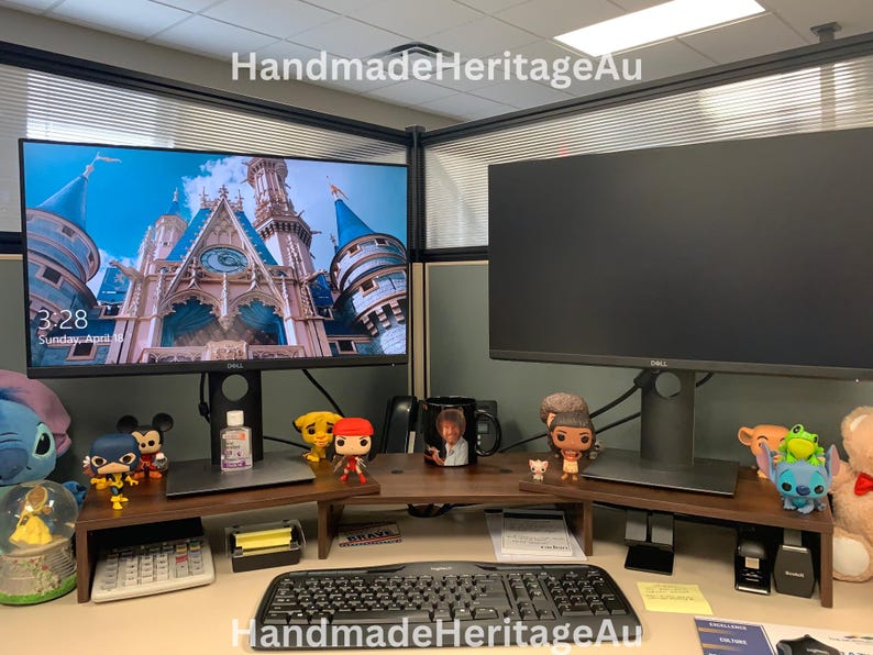 May include: A desk setup with two computer monitors, a keyboard, and decorative items. The left monitor shows a castle image. The desk includes a wooden monitor stand and figurines, including Disney characters.