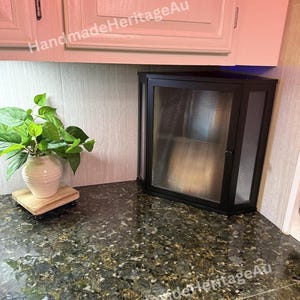 May include: A black corner cabinet with a glass door and a ribbed design. The cabinet is mounted on a countertop with a granite pattern. A white vase with green leaves sits on a wooden stand.