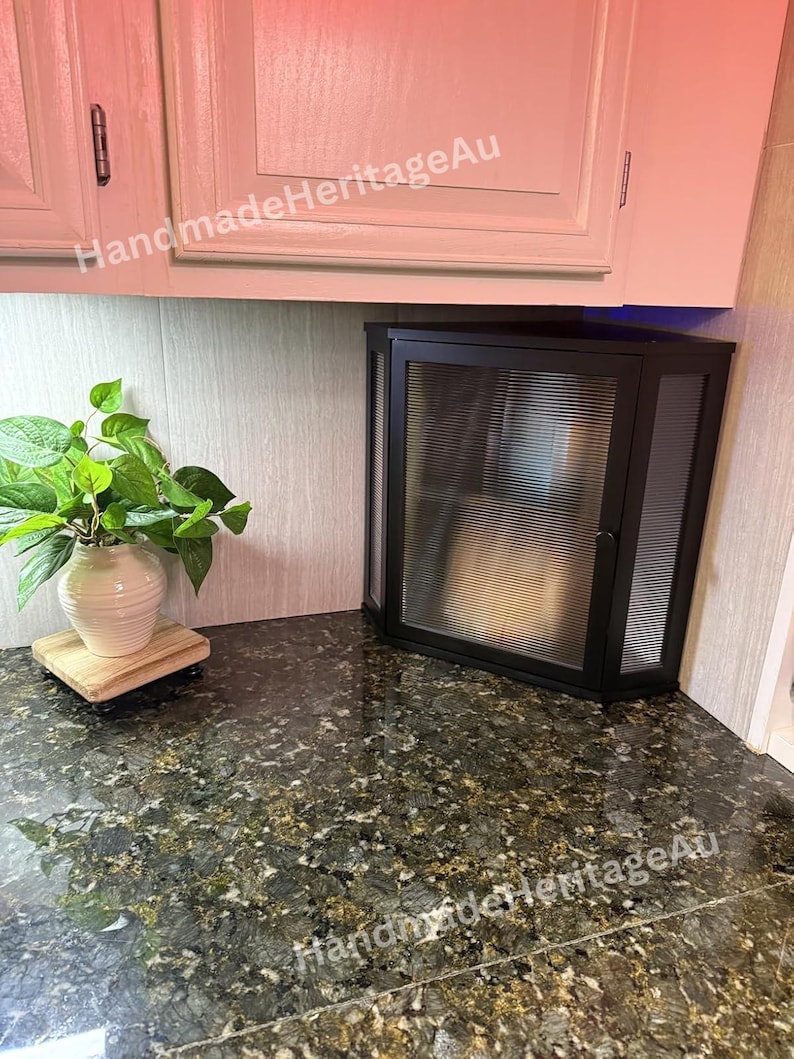 May include: A black corner cabinet with a glass door and a ribbed design. The cabinet is mounted on a countertop with a granite pattern. A white vase with green leaves sits on a wooden stand.