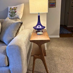 K&ouml;nnte beinhalten: Ein Beistelltisch im Mid-Century-Modern-Stil mit einer Holzplatte und einem blauen Glaslampenfu&szlig; mit cremefarbenem Lampenschirm. Auf dem Tisch steht eine Lampe. Ein dekoratives Kissen mit floralem Design liegt auf einem grauen Sofa.