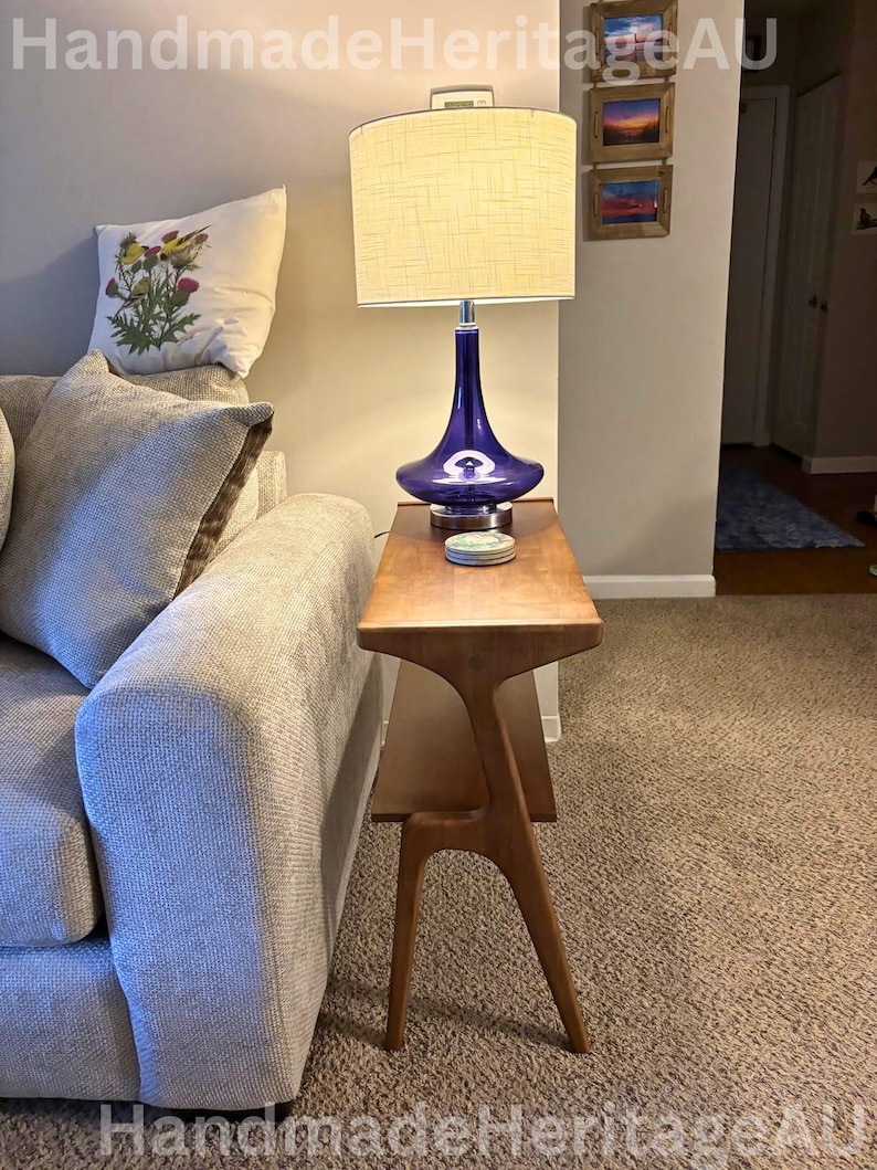K&ouml;nnte beinhalten: Ein Beistelltisch im Mid-Century-Modern-Stil mit einer Holzplatte und einem blauen Glaslampenfu&szlig; mit cremefarbenem Lampenschirm. Auf dem Tisch steht eine Lampe. Ein dekoratives Kissen mit floralem Design liegt auf einem grauen Sofa.