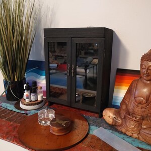 May include: A black cabinet with mirrored doors, a wooden Buddha statue, and a decorative table setting. The cabinet has ornate handles. The Buddha is brown and seated in a meditative pose. The table setting includes a wooden tray and small glass containers.