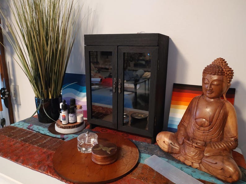 May include: A black cabinet with mirrored doors, a wooden Buddha statue, and a decorative table setting. The cabinet has ornate handles. The Buddha is brown and seated in a meditative pose. The table setting includes a wooden tray and small glass containers.