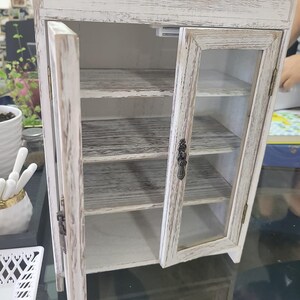 May include: A small, distressed white cabinet with glass doors and interior shelves. The cabinet has a rustic, farmhouse style, with visible wood grain and metal hardware. The doors are open, revealing the interior.