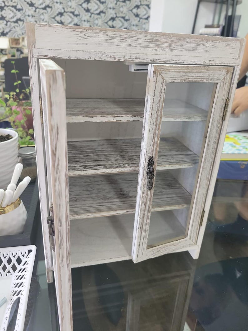 May include: A small, distressed white cabinet with glass doors and interior shelves. The cabinet has a rustic, farmhouse style, with visible wood grain and metal hardware. The doors are open, revealing the interior.