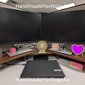 May include: A wooden desk organiser with two computer monitor stands. The stands have a dark brown finish and are angled to fit in a corner. A laptop, a small fan, and a pink heart-shaped decoration are visible.