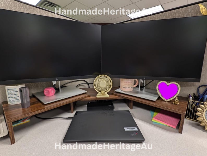 May include: A wooden desk organiser with two computer monitor stands. The stands have a dark brown finish and are angled to fit in a corner. A laptop, a small fan, and a pink heart-shaped decoration are visible.