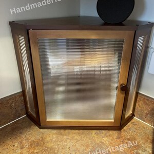 May include: A corner cabinet with a wooden frame and a ribbed glass door. The cabinet is brown with a dark top and a round black object on top. The text "HandmadeHeritageAu" is visible.