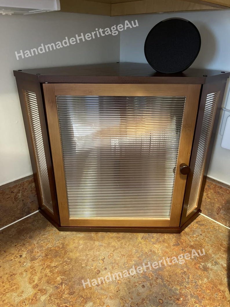 May include: A corner cabinet with a wooden frame and a ribbed glass door. The cabinet is brown with a dark top and a round black object on top. The text "HandmadeHeritageAu" is visible.