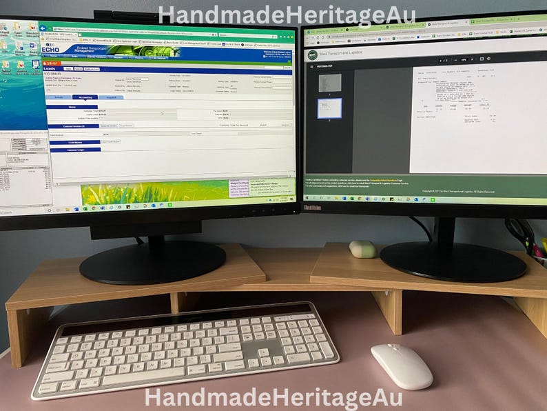 May include: A dual monitor setup with a light wood-tone monitor stand. A white keyboard and mouse are on the desk. The monitors display website content. The text "HandmadeHeritageAu" is at the bottom of the image.