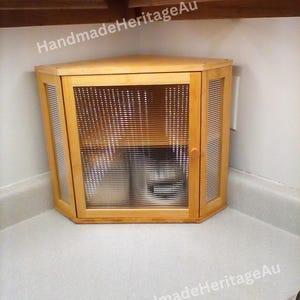 May include: A light-colored wooden corner cabinet with a glass door and slatted sides. The cabinet is positioned in a corner, with the text "HandmadeHeritageAu" visible.