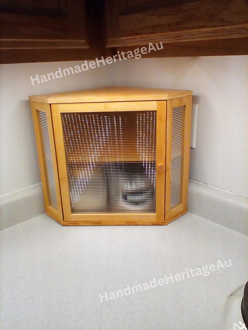 May include: A light-colored wooden corner cabinet with a glass door and slatted sides. The cabinet is positioned in a corner, with the text "HandmadeHeritageAu" visible.