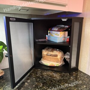 May include: A black cabinet with a ribbed glass door is open, revealing shelves stocked with food items. The cabinet is mounted under a white cabinet. The countertop is a dark granite with gold flecks. The text "HandmadeHeritageAu" is visible.