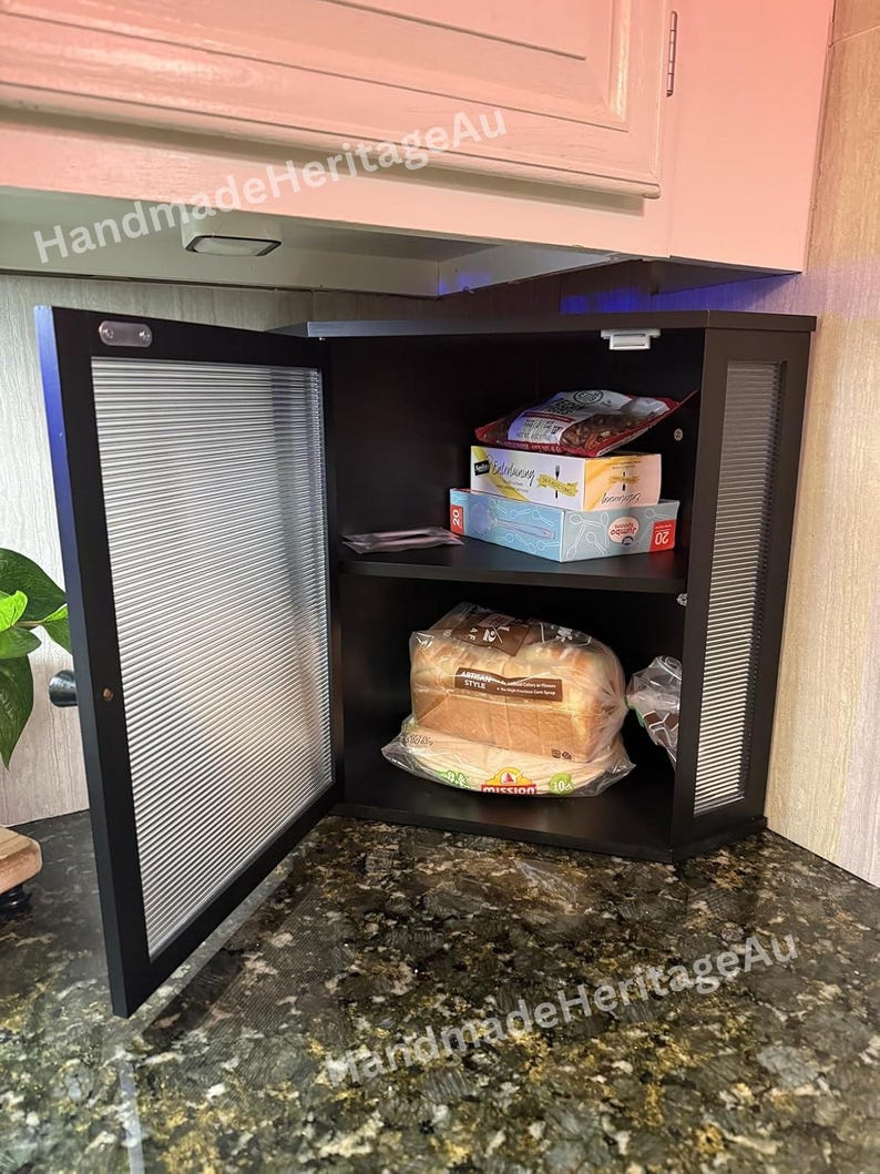 May include: A black cabinet with a ribbed glass door is open, revealing shelves stocked with food items. The cabinet is mounted under a white cabinet. The countertop is a dark granite with gold flecks. The text "HandmadeHeritageAu" is visible.
