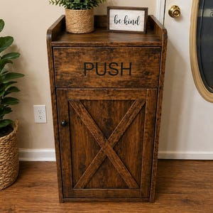 May include: A wooden cabinet with a dark brown finish. The cabinet has a drawer with the word "PUSH" printed on it and a door with an X-shaped design. A small sign that says "be kind" sits on top, along with a potted plant.