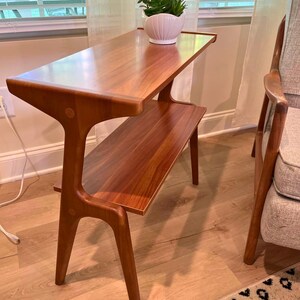 K&ouml;nnte beinhalten: Ein zweistufiger Beistelltisch im Mid-Century-Modern-Stil mit warmem, rotbraunem Holzfinish. Die obere Ebene ist rechteckig, die untere etwas kleiner. Auf der Oberseite steht ein wei&szlig;er Keramiktopf mit einer Pflanze.