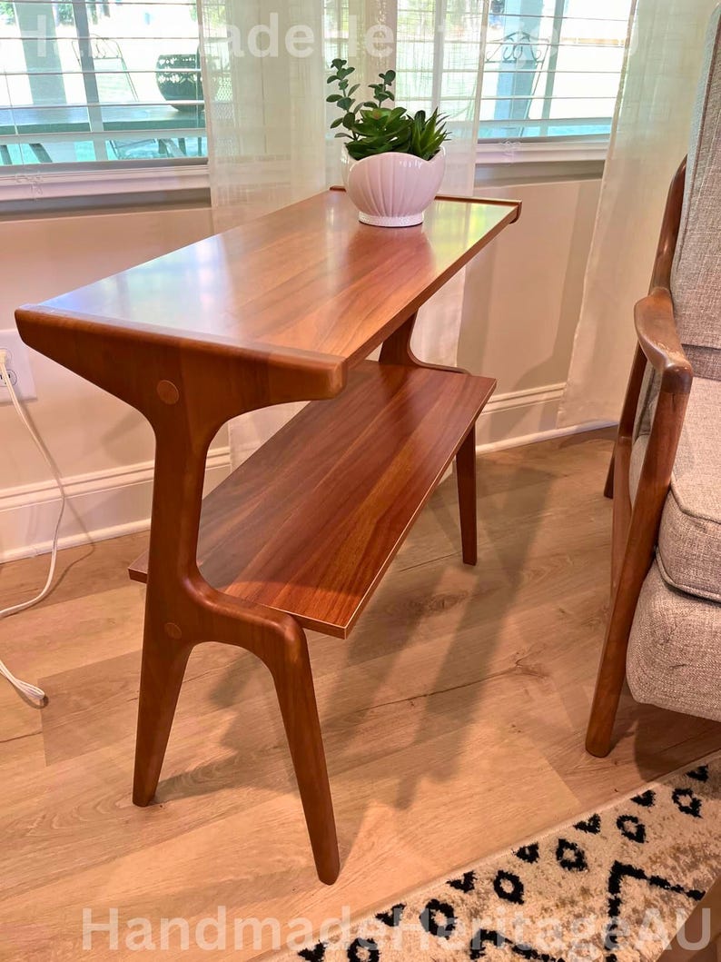 K&ouml;nnte beinhalten: Ein zweistufiger Beistelltisch im Mid-Century-Modern-Stil mit warmem, rotbraunem Holzfinish. Die obere Ebene ist rechteckig, die untere etwas kleiner. Auf der Oberseite steht ein wei&szlig;er Keramiktopf mit einer Pflanze.