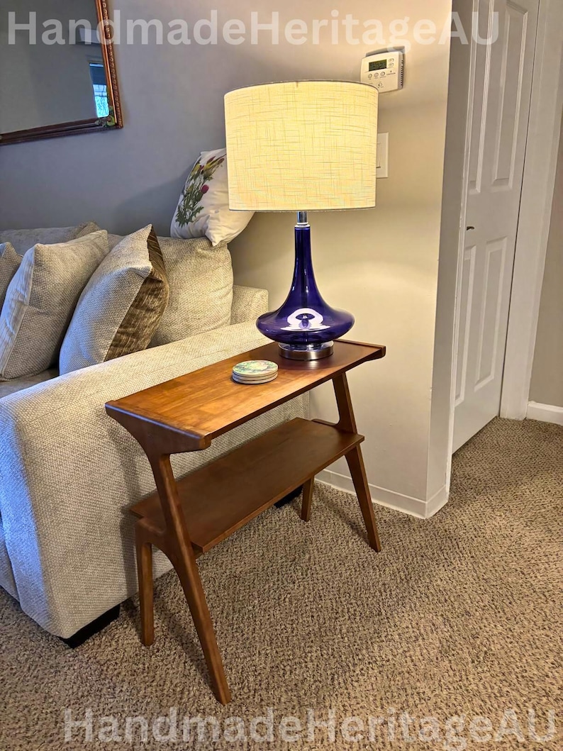 K&ouml;nnte beinhalten: Ein Beistelltisch im Mid-Century-Modern-Stil mit zwei Ebenen, aus warm get&ouml;ntem Holz gefertigt. Eine blaue Glaslampe mit cremefarbenem Lampenschirm steht auf dem Tisch. Die Lampe hat einen einzigartigen, geschwungenen Sockel. Der Tisch steht neben einem Sofa.