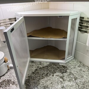 May include: White corner cabinet with a door ajar, revealing two shelves. The cabinet has a textured glass panel on the side and a slatted door. The interior contains brown paper. The cabinet is set on a speckled countertop.