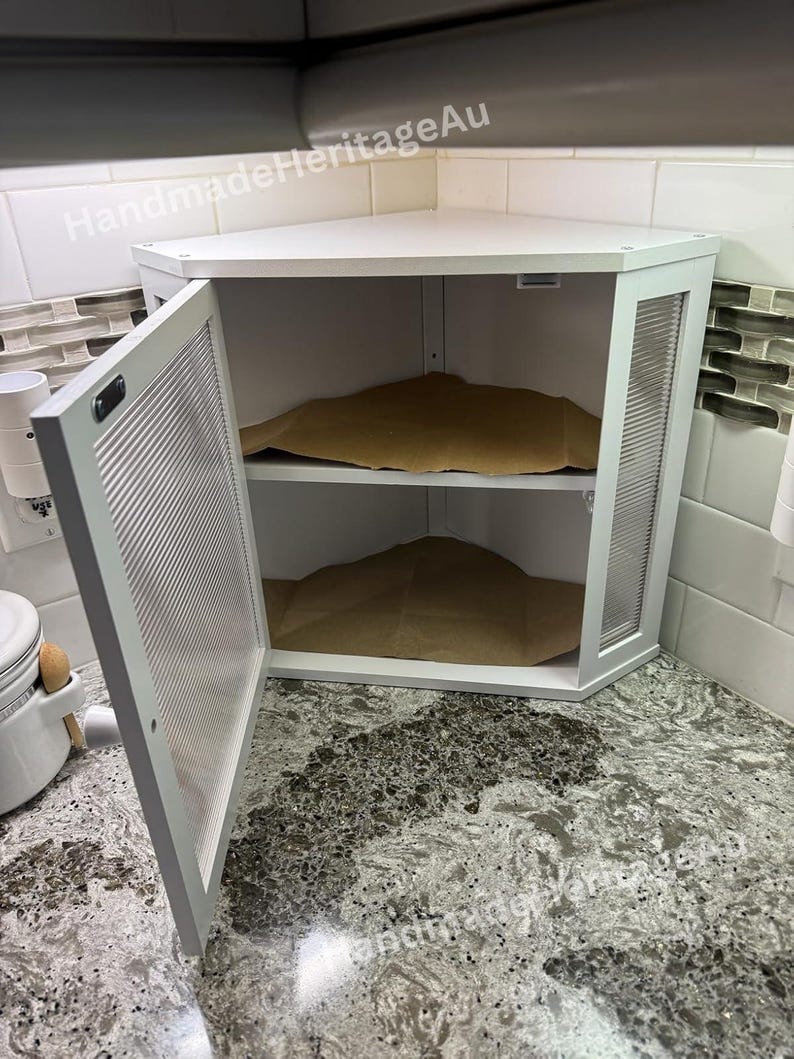 May include: White corner cabinet with a door ajar, revealing two shelves. The cabinet has a textured glass panel on the side and a slatted door. The interior contains brown paper. The cabinet is set on a speckled countertop.