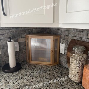 May include: A light wood corner cabinet with a ribbed glass door is installed in a kitchen. The cabinet is surrounded by white cabinets and a gray brick backsplash. A paper towel holder and jars of food are also visible.