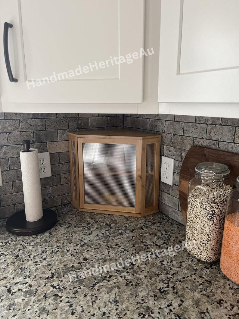 May include: A light wood corner cabinet with a ribbed glass door is installed in a kitchen. The cabinet is surrounded by white cabinets and a gray brick backsplash. A paper towel holder and jars of food are also visible.