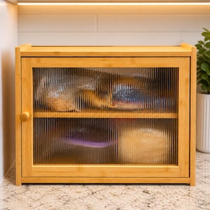 May include: A light-colored bamboo cabinet with a glass door. The cabinet has two shelves and a round wooden knob. The glass door has a vertical ribbed texture. The cabinet is on a countertop.