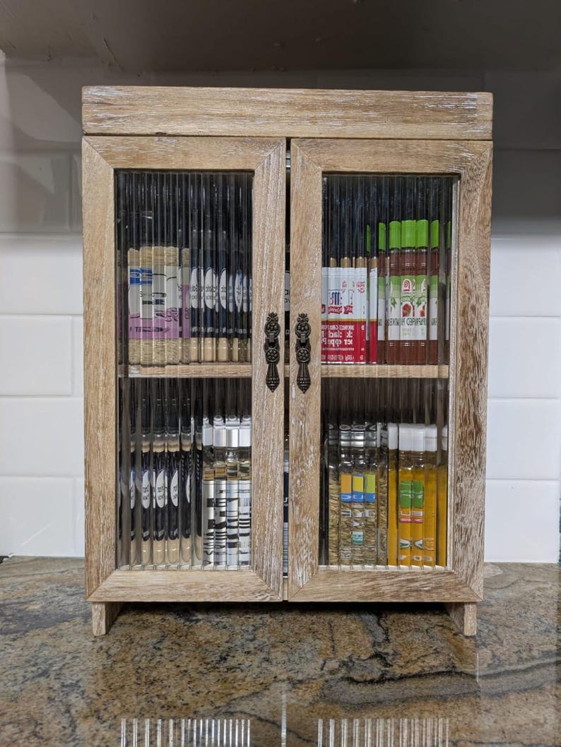 May include: A wooden cabinet with glass doors, featuring a variety of glass bottles and jars, some with labels. The cabinet has a distressed white finish and is ideal for storing and displaying small items.