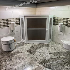 May include: A white corner cabinet with a glass door and a handle is installed in a kitchen. The cabinet is surrounded by white tiles and a speckled countertop. Two electrical outlets are visible on the wall.