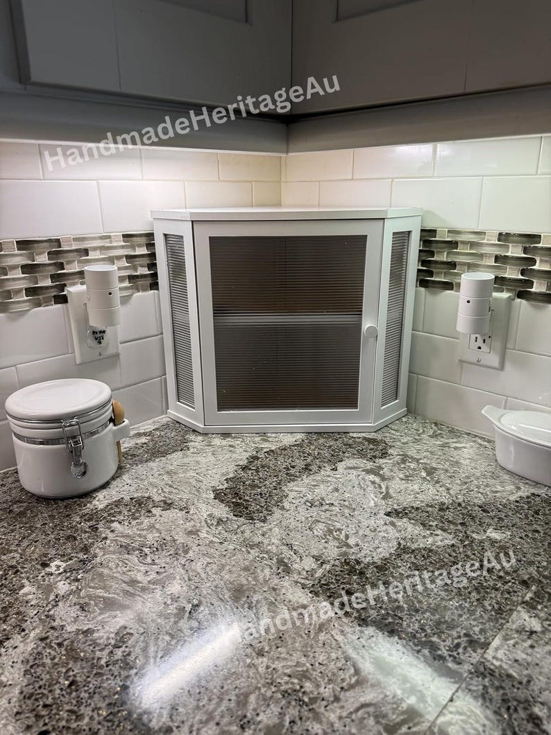 May include: A white corner cabinet with a glass door and a handle is installed in a kitchen. The cabinet is surrounded by white tiles and a speckled countertop. Two electrical outlets are visible on the wall.