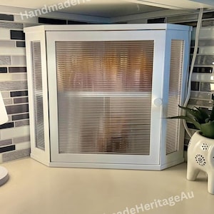 May include: White corner cabinet with a ribbed glass door and side panels. The cabinet has a small handle and is mounted on a countertop. The text "HandmadeHeritageAu" is visible on the top and bottom of the cabinet.